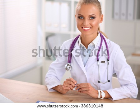 Beautiful young smiling female doctor sitting at the desk and writing.