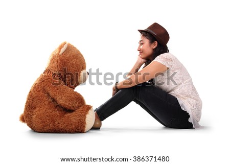Teenage girl in hipster style sits facing her brown teddy bear on the floor, as if they're having a conversation, isolated on white background