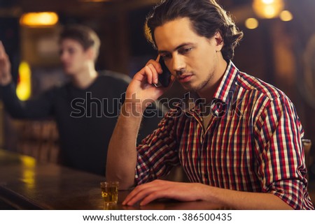 Sad attractive man in casual clothes is talking on the mobile phone while sitting at bar counter in pub