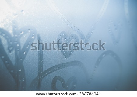 Closeup on abstract blurred love heart symbols drawn by hand on the wet frozen window glass sunlight background. Selective focus used.