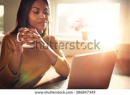 Adult woman smiling sitting near bright window while looking at open laptop computer on table and holding white mug