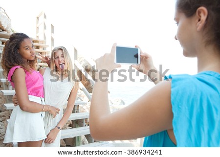 Joyful diverse teenagers girls friends pulling faces and posing together for photos with smart phone, sunny coastal outdoors. Adolescents summer travel lifestyle. Young people using technology.