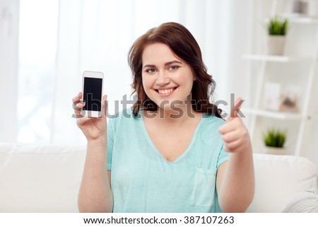 people, technology, communication and leisure concept - happy young plus size woman sitting on sofa and showing smartphone blank screen and showing thumbs up at home