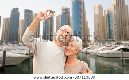 age, tourism, travel, technology and people concept - senior couple with camera taking selfie over dubai city harbor or waterfront background