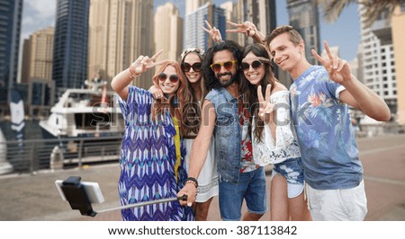 nature, summer, youth culture, technology and people concept - smiling hippie friends in sunglasses taking picture by smartphone on selfie stick and showing peace gesture over dubai city harbour 