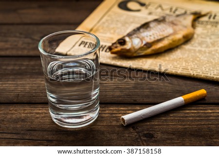 Shot of vodka with a stockfish and newspaper with cigarette on an old wooden table. Close up view