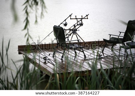Fishing tackle gear two convertible chairs stay outdoor in rain drops falling plop into water on wet wooden pier over natural rainy day background, horizontal picture