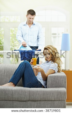 Happy woman sitting at couch watching TV, man ironing in the background. Selective focus on woman.