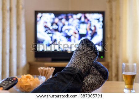 man watching American football match on TV (television) with feet on table, eating snacks - stock photo