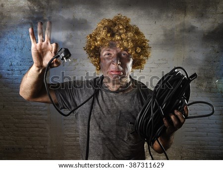 man with funny wig holding electrical cable smoking after domestic accident with dirty burnt face and shock electrocuted expression in electricity DIY repairs danger concept in black smoke background
