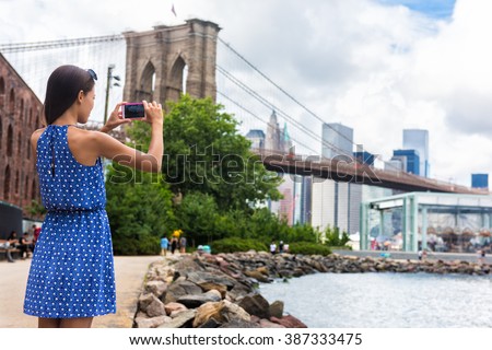 Tourist taking travel picture with phone of Brooklyn bridge and New York City skyline during summer holidays. Unrecognizable female young adult enjoying USA vacations in blue dress.