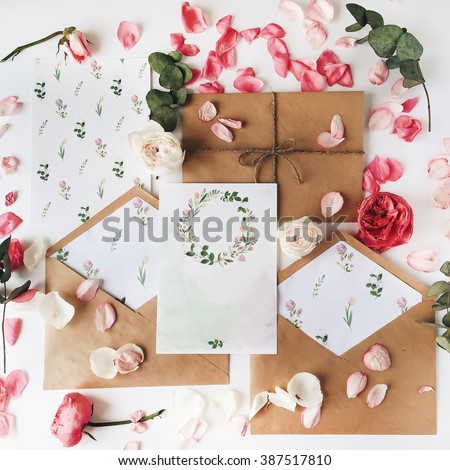 Workspace. Wedding invitation cards, craft envelopes, pink and red roses and green leaves on white background. Overhead view. Flat lay, top view Royalty-Free Stock Photo #387517810