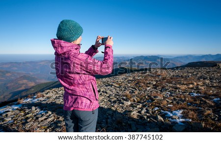 Young woman hiker taking picture with her smart phone on the peak of the rocky mountain. Rear view, high altitude. Cold and sunny fall day.