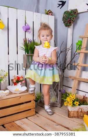Sweet girl in a light dress standing in full length with a toy chick in hand. Bright colorful spring decorations