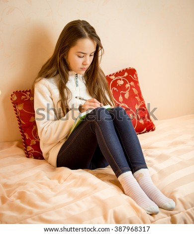 Teenage girl sitting on bed and writing poems in notebook Royalty-Free Stock Photo #387968317