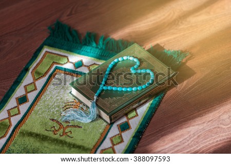 Mat for prayer , the rosary and the holy book the Koran. On the cover of the book in Arabic is written the name of the book (translated ) - Quran . Royalty-Free Stock Photo #388097593