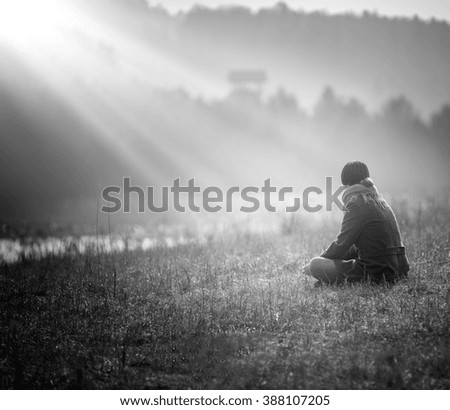 Young woman sitting on grass on meadow near lake and dreaming. Photo taken from behind.