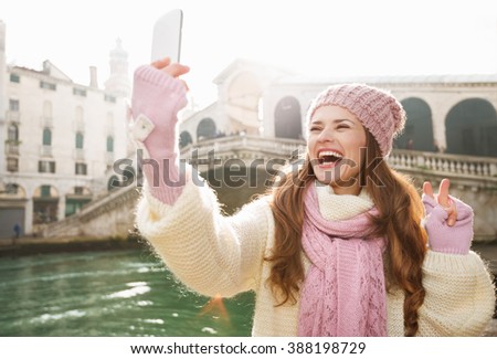 Ultimate getaway shortcut - start New Year going on Carnival in Venice, Italy. Happy woman tourist in white sweater showing victory while taking selfie with smartphone in the front of Rialto Bridge