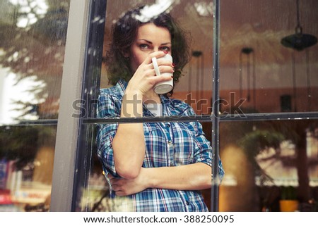 beautiful young woman standing near window, drinking the coffee and looking into the distance