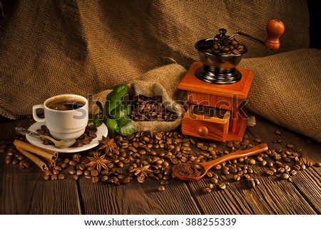 Coffee beans in jute bag with coffee grinder, hot coffee cup, cinnamon sticks, anise and green leaves on a wooden table. Royalty-Free Stock Photo #388255339
