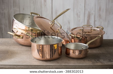 A set of copper cookware that include a small and medium-sized saucepan, a large saucepan, pot, and cover stacked on top of the other, and a covered small pot, all placed on a wooden countertop. Royalty-Free Stock Photo #388302013