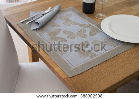 A rectangular placemat with embroidered butterflies as design spread out on wooden table with napkin and utensils atop alongside empty dinner plate, wine glass, and a bottle of wine. Royalty-Free Stock Photo #388302508