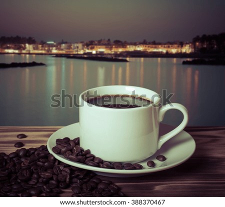 Coffee beans and coffee in white cup on wooden table opposite a defocused natural landscape for background. Collage. Selective focus. Toned.