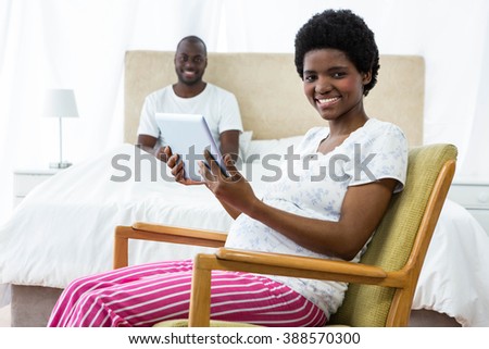 Portrait of pregnant woman using digital tablet on chair and man sitting on bed in background