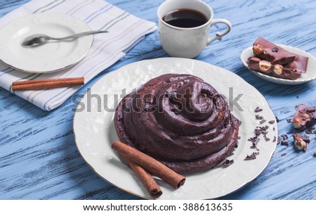 Round Twisted Bun Danish snail poured chocolate icing, white cup of coffee espresso, cinnamon sticks, pieces of chocolate with hazelnuts on a blue wooden background