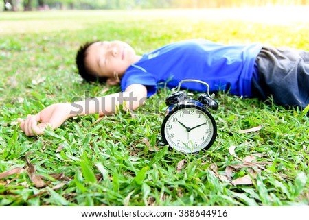 Selective focus on the classical black alarm clock model, in front of the sleeping young boy on green lawn in the park in day time.
