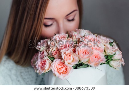 Closeup portrait Nice young blonde girl with Blue Eyes and healthy Long Blond Hair holding roses bouquet in hat box  against the  plastered wall, wearing jeans and knit sweater, smelling flowers. Royalty-Free Stock Photo #388673539