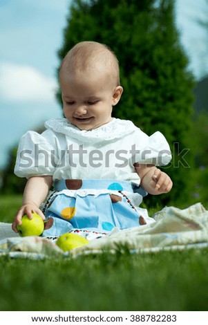Happy smiling funny small baby girl in stylish dress sitting outdoor on picnic with green apples on natural grass backdrop, vertical picture