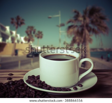 Coffee beans and coffee in white cup on wooden table opposite a defocused urban landscape for background. Collage. Selective focus. Toned.