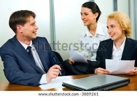 Portrait of two businesswomen and businessman sitting at the table with closed laptop holding documents and discussing questions