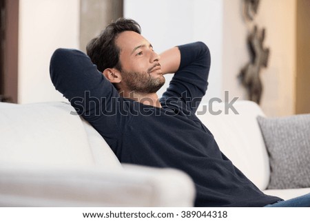 Portrait of a young man resting on sofa and thinking about the future. Handsome young man with hands behind head sitting on couch in living room. Positive man daydreaming and relaxing at home. 
