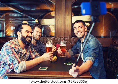 people, men, leisure, friendship and technology concept - happy male friends drinking beer and taking picture with smartphone selfie stick at bar or pub