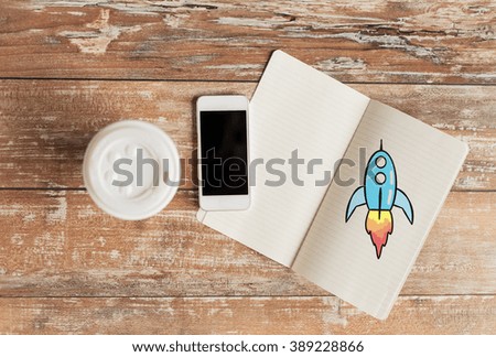 business, education, objects and technology concept - close up of coffee paper cup, smartphone and rocket drawing in notebook on table