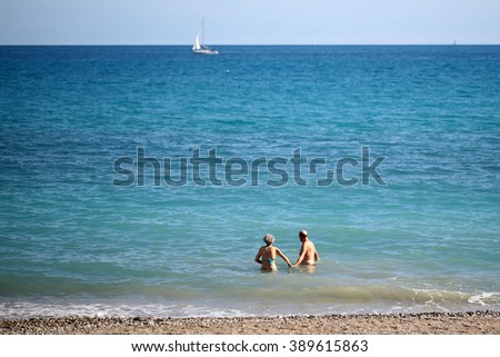 Photo closeup of senior couple woman man holding hands goes for dip into sea with waves running pebble beach at seashore and yacht sailing boat offshore on blurred blue background, horizontal picture