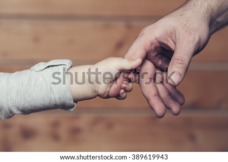 Closeup of two touching hands of small baby boy holding finger of male father as symbol of family love and trust on blurred wooden background, horizontal picture