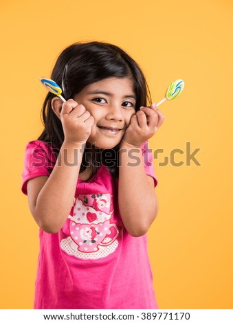 Adorable little Indian/asian girl eating Lollipop or Sweet Candy with happy expressions. Standing isolated over yellow background