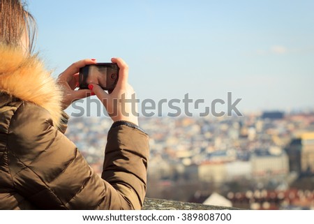 Pretty young girl taking / shooting a picture / video of big history city during the winter sunset (focus on camera; color toned image)