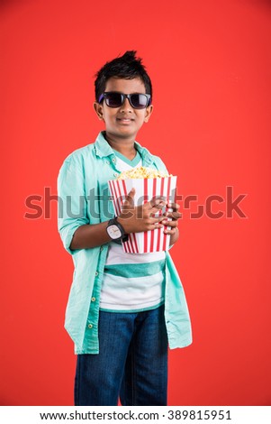 Cute happy little Indian boy eating popcorn  from box while wearing sunglass or 3D glasses in a theatre. Standing isolated over colourful background