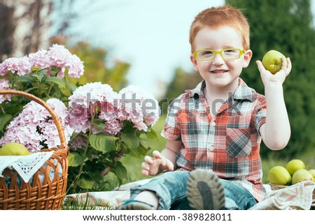 Happy smiling funny small red haired boy in stylish checkered shirt jeans and yellow glasses sitting outdoor on picnic with basket of green apples near flowers on natural backdrop, horizontal picture