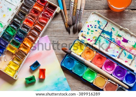 Set of watercolor paints, brushes for painting and paper sheet of painting on old wooden background. Top view.