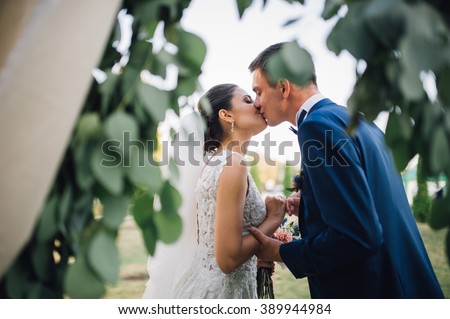couple in wedding attire exchange rings with a bouquet of flowers and greenery in the garden with arch on background, the bride and groom Royalty-Free Stock Photo #389944984