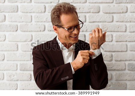Handsome middle aged businessman in classic suit and eyeglasses is adjusting a cufflink and smiling, standing against white brick wall