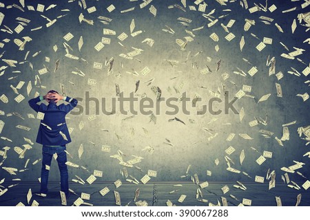 Rear back view of business man standing in front of a wall under money rain dollar banknotes falling down, hands on head wondering what to do next. Full body length of businessman facing the wall