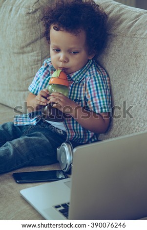 Watching cartoons online. High angle view of little African baby boy drinking from baby bottle and looking at his laptop while sitting on the couch at home 