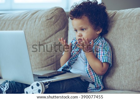 How does this thing turns on? Little African baby boy looking confused while sitting on the couch at home with laptop on his knees Royalty-Free Stock Photo #390077455