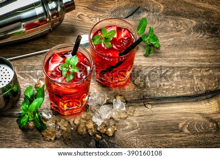 Glasses of red cocktail with ice, mint leaves and strawberry on wooden kitchen table. Vintage style toned picture
